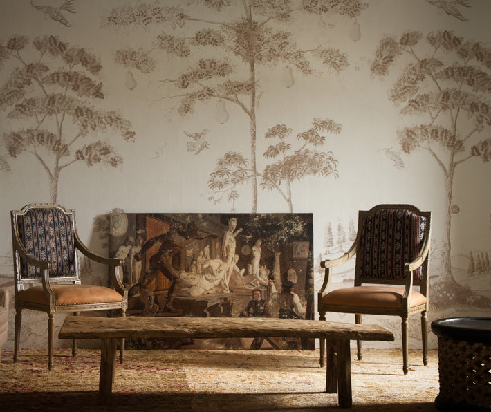 Lifestyle shot of Mythical Land wallpaper, in clay on a saturated white background and sepia tones to illustrate the lively river bordered with wild flowers, trees growing giant pears and a whole dance of magical creatures, with antique furniture in the foreground.