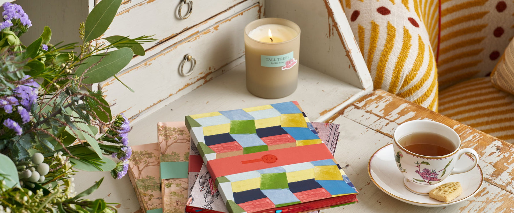 A stack of colourful notebooks sit on an antique desk with a cup of tea, a candle and a vase of purple, white and green flowers