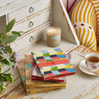 A stack of colourful notebooks sit on an antique desk with a cup of tea, a candle and a vase of purple, white and green flowers