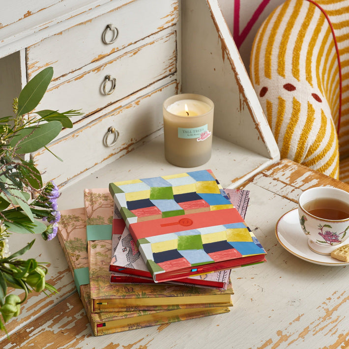 A stack of colourful notebooks sit on an antique desk with a cup of tea, a candle and a vase of purple, white and green flowers