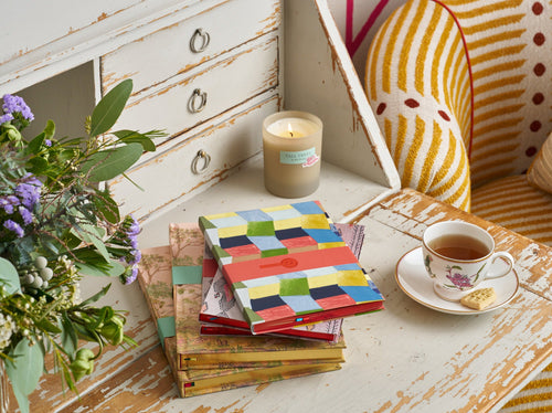 A stack of colourful notebooks sit on an antique desk with a cup of tea, a candle and a vase of purple, white and green flowers