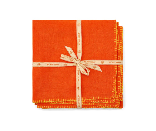 A stack of orange linen napkins with yellow blanket stitch , tied with canvas shop Kit Kemp ribbon.