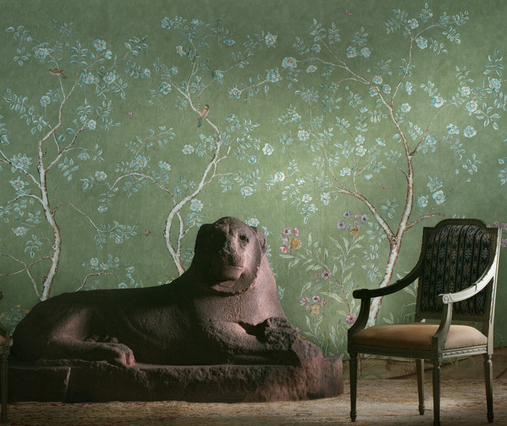 Kit's New Songbird Wallpaper has a the stylised nature of Chinoiserie landscapes with an English twist, in the Spring colourway suffused in the soft green background, where a breeze ruffles the leaves of loosely painted trees dotted with dog roses, shown with large lion sculpture in brown, and upholstered Louis 15th chair.