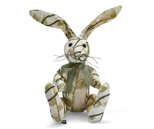 The image shows a stuffed toy rabbit made from patterned fabric. The rabbit has long ears and is sitting upright. It appears to be handmade, with visible stitching and a slightly patchwork look from the different fabric patterns used. The rabbit's body is primarily covered in a cream patterned fabric that resembles a woven texture featuring leaves and vines. 