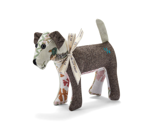 The image depicts a handmade, patchwork toy dog. The dog is made of a combination of brown and patterned fabrics, with a ribbon tied around its neck. The dog features an embroidered duck on the side of its face.