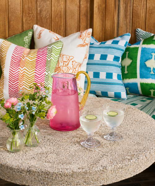 Colorful pillows on a bench with a pink pitcher, glasses, and flowers on a stone surface.