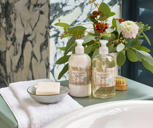 Life style image of Tall trees shampoo and conditioner, presented beside roll top bath, on a sage green table.