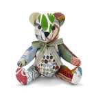 The image depicts a plush toy in the shape of a teddy bear. The bear is made up of various patterned and textured fabrics, including stripes, polka dots, and abstract designs in a range of colours such as green, brown, and blue. The bear is wearing a ribbon around its neck with the text Firmdale Hotels printed on it.