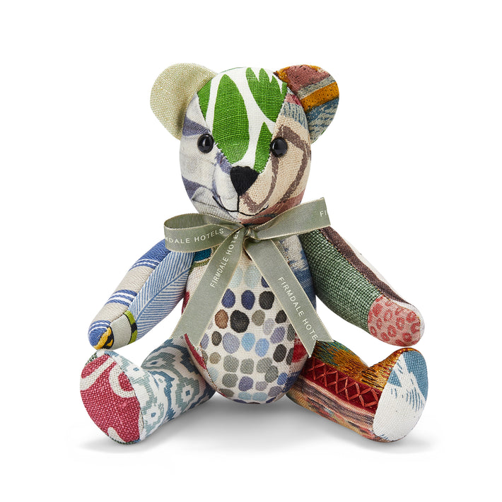 The image depicts a plush toy in the shape of a teddy bear. The bear is made up of various patterned and textured fabrics, including stripes, polka dots, and abstract designs in a range of colours such as green, brown, and blue. The bear is wearing a ribbon around its neck with the text Firmdale Hotels printed on it.