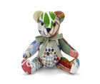 The image depicts a plush toy in the shape of a teddy bear. The bear is made up of various patterned and textured fabrics, including stripes, polka dots, and abstract designs in a range of colours such as green, brown, and blue. The bear is wearing a ribbon around its neck with the text Firmdale Hotels printed on it.